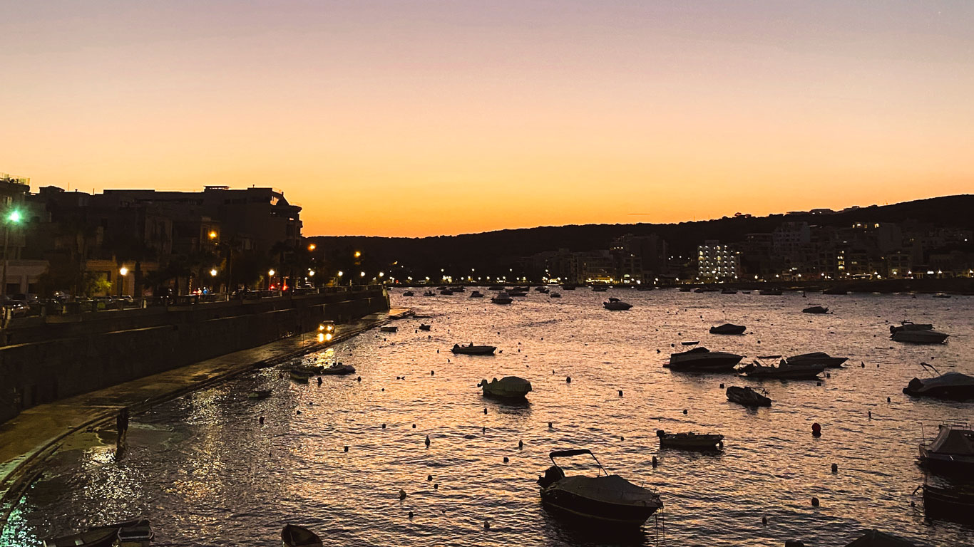 El crepúsculo cae sobre la bahía de San Pablo en Malta, con los últimos tonos del atardecer lanzando un resplandor cálido sobre el agua, donde los barcos se balancean suavemente y el paseo marítimo cobra vida con las luces de la noche.