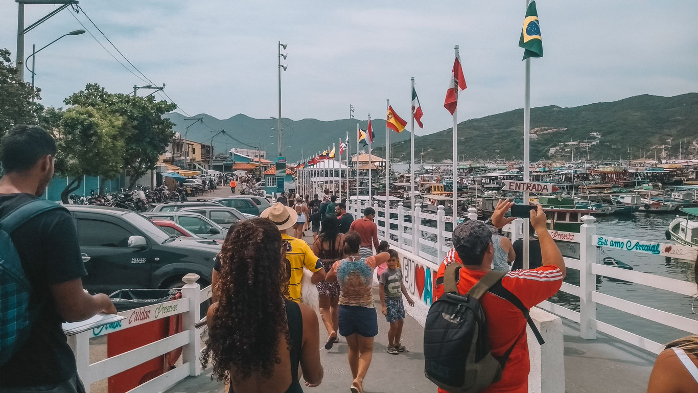 Turistas caminan por el muelle de la Marina dos Pescadores en Arraial do Cabo, con una hilera de banderas ondeando y barcos anclados en el fondo, en un día animado con cielo nublado sobre montañas verdes.