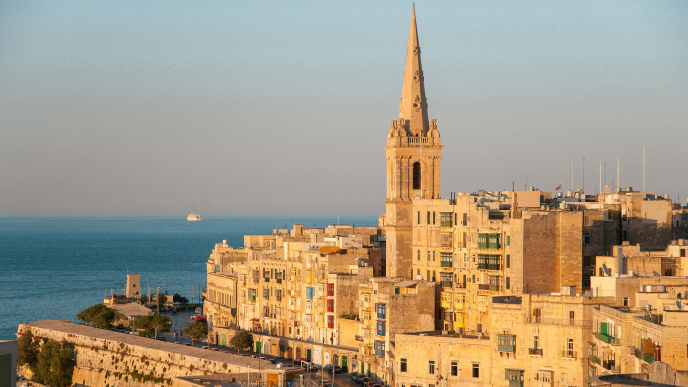 Vista iluminada por el sol de la Valletta en Malta, mostrando los históricos edificios de piedra arenisca con sus tradicionales balcones y un llamativo campanario de iglesia que domina el horizonte, todo ello contra el telón de fondo del mar Mediterráneo.