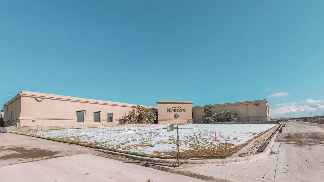 Vista exterior de Bodega Norton en Mendoza, Argentina. El gran edificio beige con el nombre de la bodega prominentemente mostrado en una pared central se encuentra bajo un cielo azul claro. La nieve cubre el suelo circundante y las cumbres montañosas distantes, añadiendo al apacible paisaje invernal.