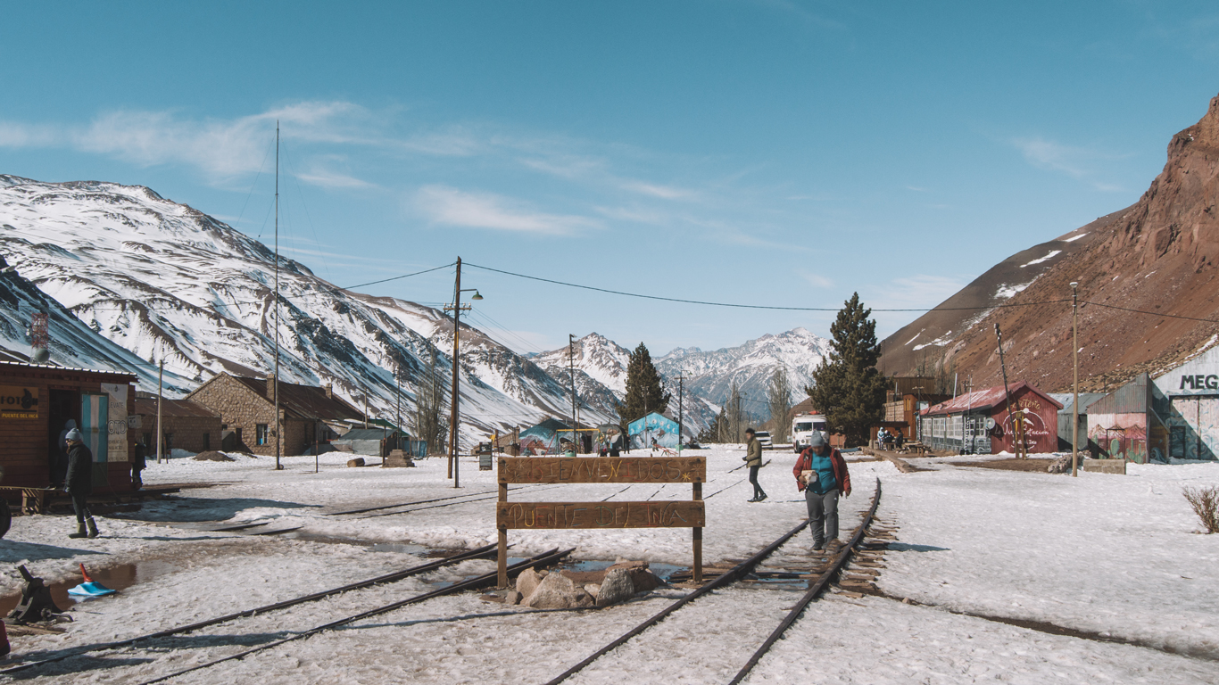 Una pequeña aldea nevada en Mendoza, con casas de madera y montañas cubiertas de nieve al fondo. La escena muestra vías de tren que cruzan el pueblo, donde algunos visitantes disfrutan del paisaje andino en un día soleado de agosto.