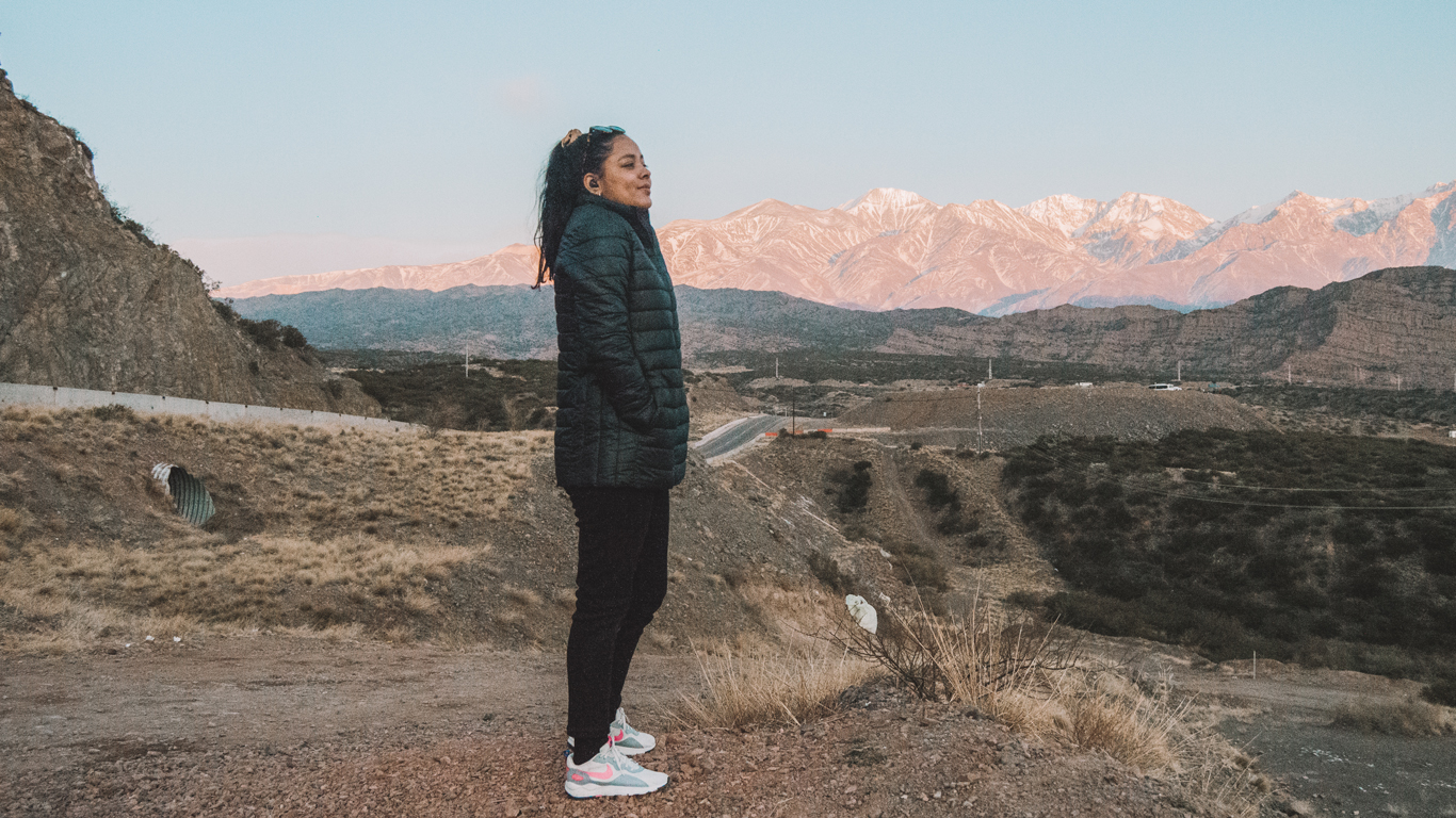 Una mujer con una chaqueta de invierno está de pie en un paisaje árido con montañas nevadas al fondo, iluminadas por la luz del amanecer o atardecer. La imagen captura la belleza de Mendoza en agosto, mostrando un contraste entre el terreno seco y las cumbres nevadas de los Andes.