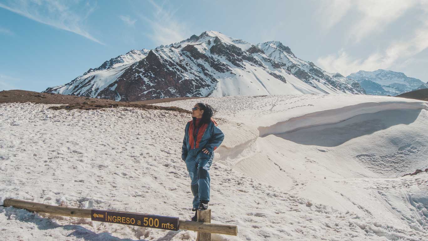 Una mujer en ropa de invierno está de pie sobre una señal que indica "INGRESO a 500 mts." en un paisaje nevado con montañas en el fondo. La escena se desarrolla en Mendoza, Argentina, mostrando la belleza de los Andes nevados.