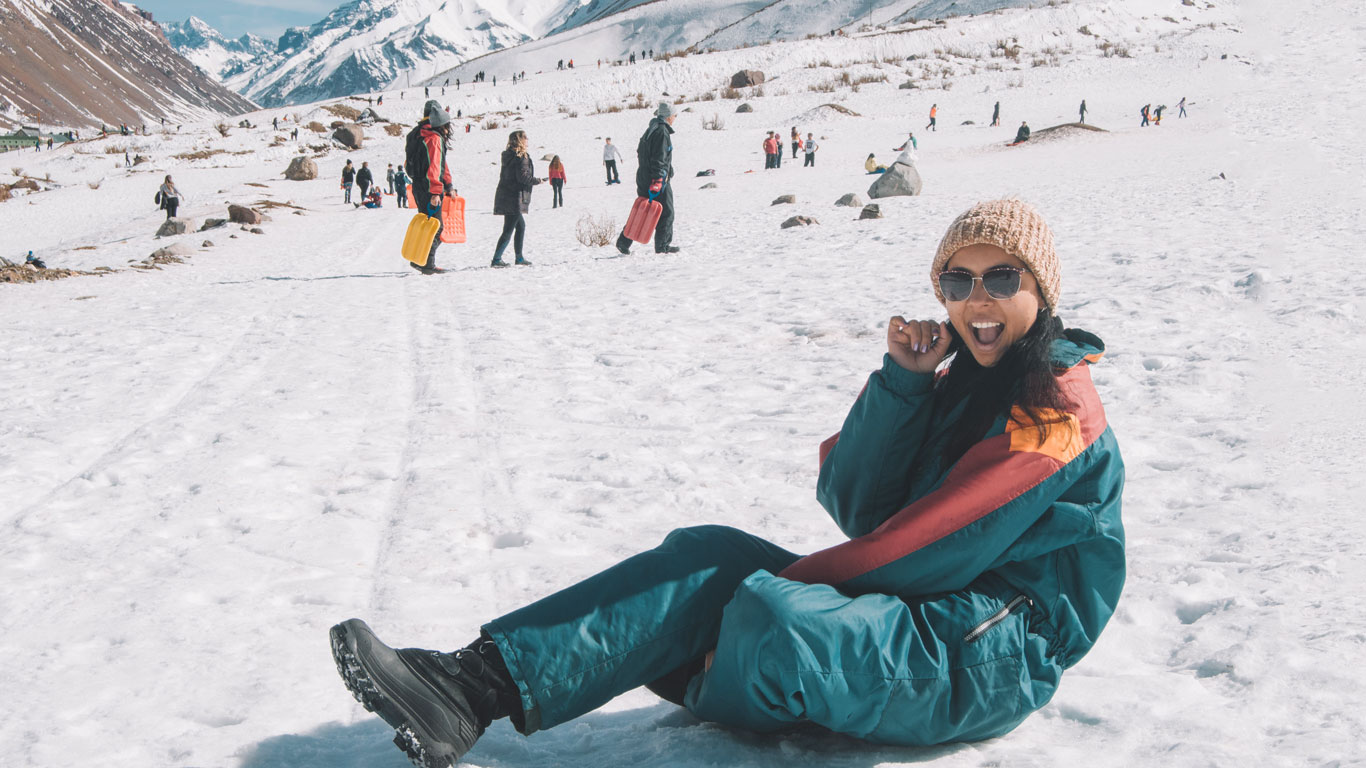 Una mujer alegre sentada en una pendiente nevada en Mendoza, Argentina, vistiendo una chaqueta de invierno colorida, gafas de sol y un gorro de punto beige. Sonríe ampliamente a la cámara, disfrutando de las actividades invernales a su alrededor. El fondo presenta una escena bulliciosa de otros visitantes participando en juegos de nieve, con montañas escarpadas que se extienden hasta el horizonte.