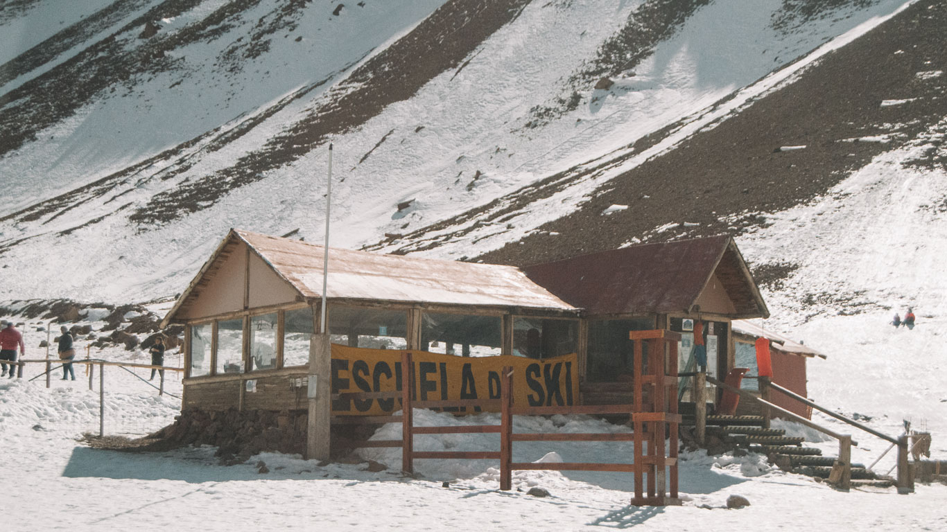 Fotografía de una escuela de esquí en un centro turístico de montaña en Mendoza durante julio. La estructura de madera, etiquetada como 'ESCUELA DE SKI', se encuentra en la base de una montaña parcialmente cubierta de nieve. Se pueden ver personas alrededor del edificio, disfrutando del entorno invernal bajo un cielo despejado.