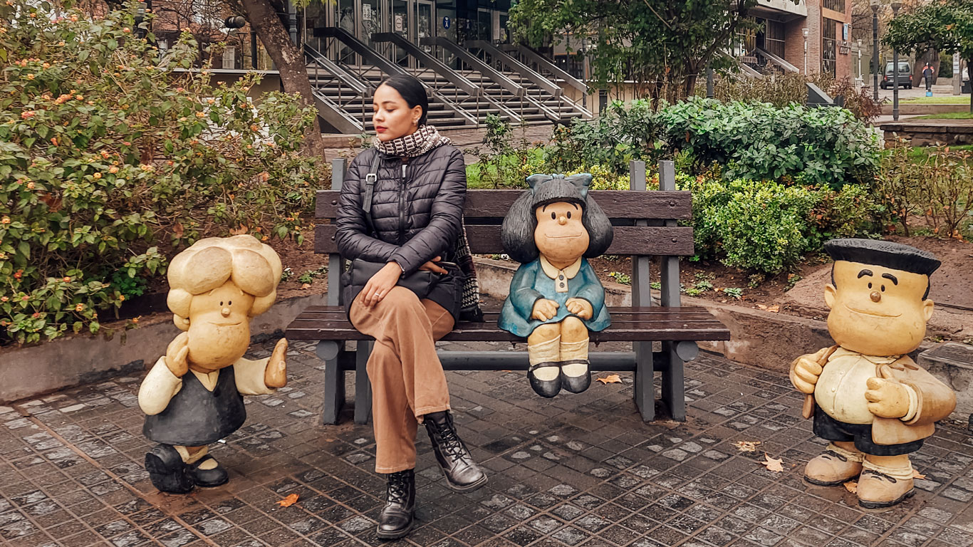 Una mujer se sienta en un banco del parque entre estatuas de dibujos animados coloridas en Mendoza, Argentina. A su izquierda hay una estatua de un niño con un peinado grande y zapatos grandes, y a su derecha, una niña con el pelo negro largo y un vestido azul. Ambas estatuas parecen alegres y están estilizadas para parecerse a personajes de libros infantiles, añadiendo un toque caprichoso al entorno urbano.