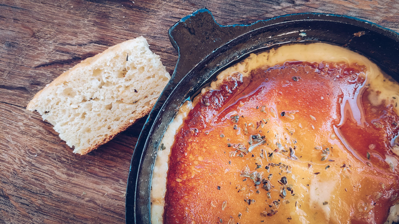 Un plato de provoleta dorada, cubierta con orégano y acompañada de una rebanada de pan, sobre una mesa de madera. La provoleta es un tradicional queso provolone argentino, servido derretido y especiado, representando una delicia culinaria de Argentina.