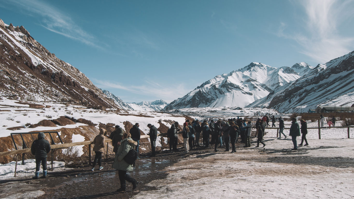 Un grupo de personas abrigadas camina sobre un sendero nevado en Puente del Inca, Mendoza, con las majestuosas montañas de los Andes de fondo. La escena destaca la popularidad del destino turístico y la belleza invernal del paisaje en agosto.