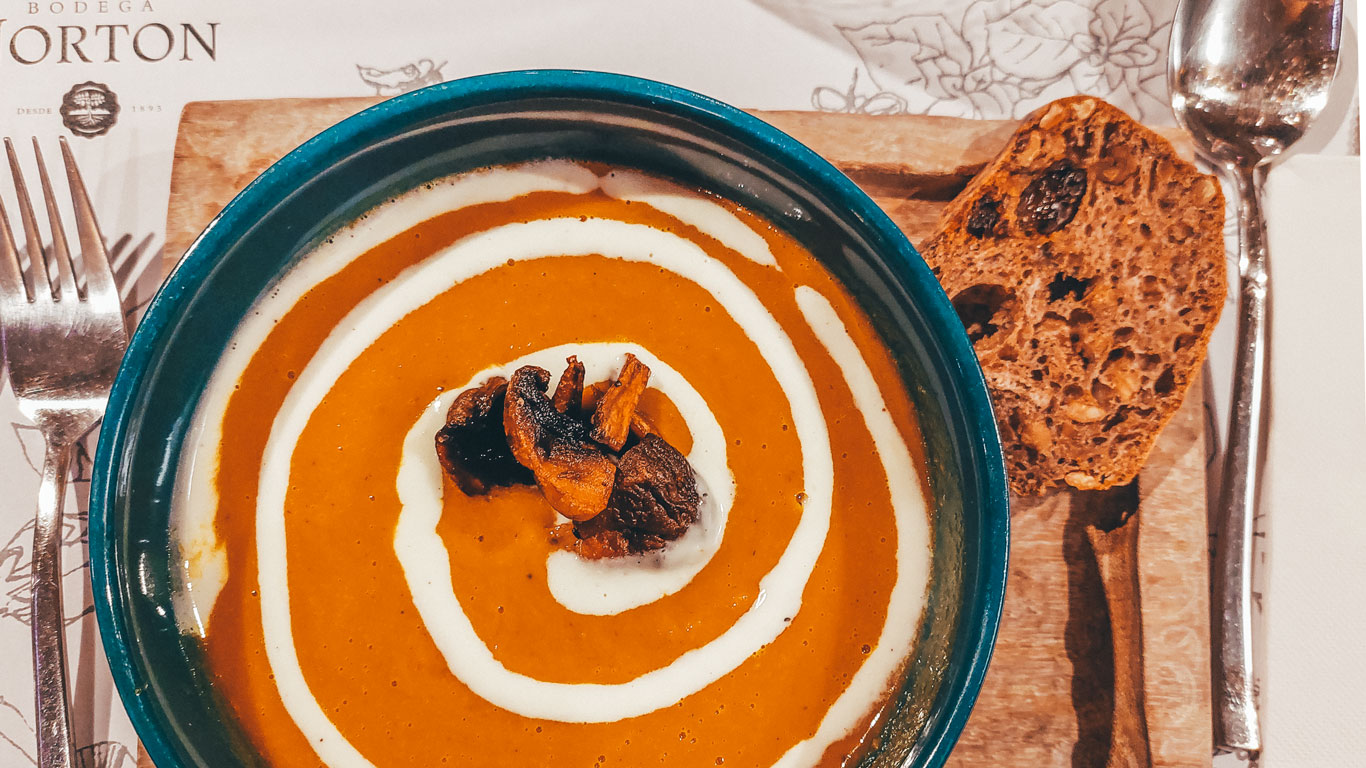 Sopa de zanahoria naranja vibrante servida en el Restaurante La Vid, Bodega Norton, Mendoza. La sopa está elegantemente adornada con un remolino de crema y champiñones y semillas de calabaza asados en el centro, acompañada de una rebanada de pan multicereal en una tabla de madera. La mesa está dispuesta con cubiertos y una servilleta, todo bajo una luz suave de comedor.