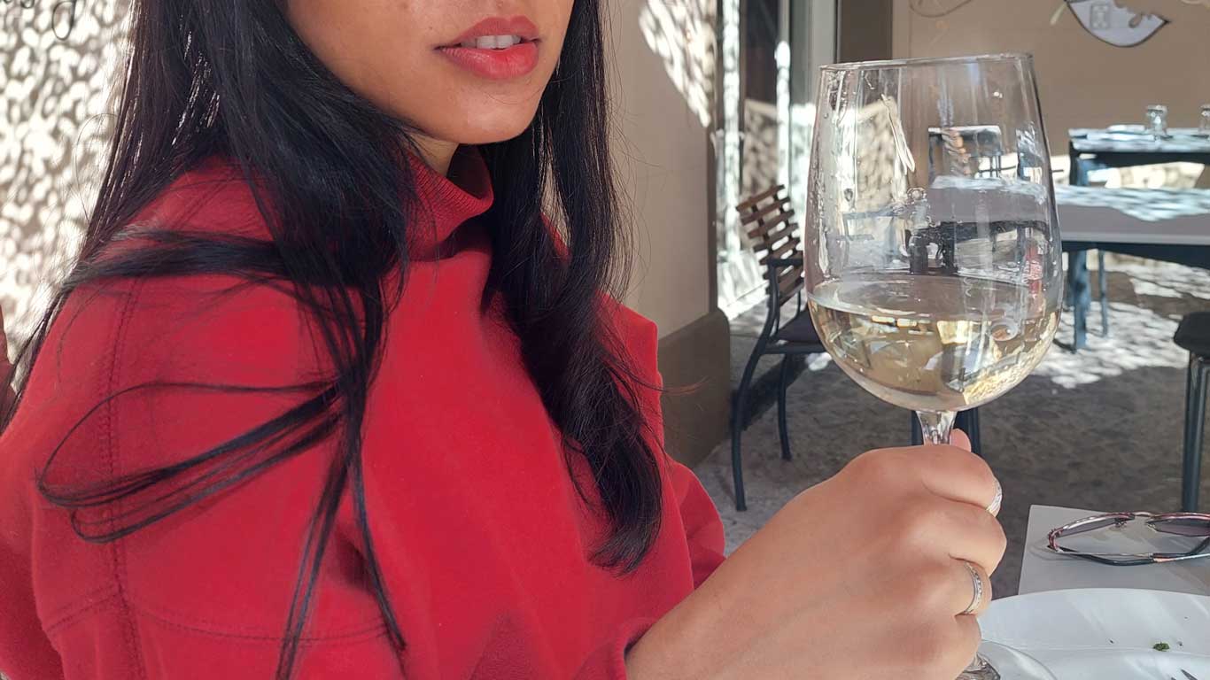 La fotografía muestra a una mujer disfrutando de una copa de vino blanco en una terraza soleada, capturando la esencia de una experiencia enológica. Ella lleva una chaqueta roja vibrante y sostiene delicadamente la copa, observando el vino con aprecio mientras la luz natural acentúa el brillante color ámbar de la bebida. 