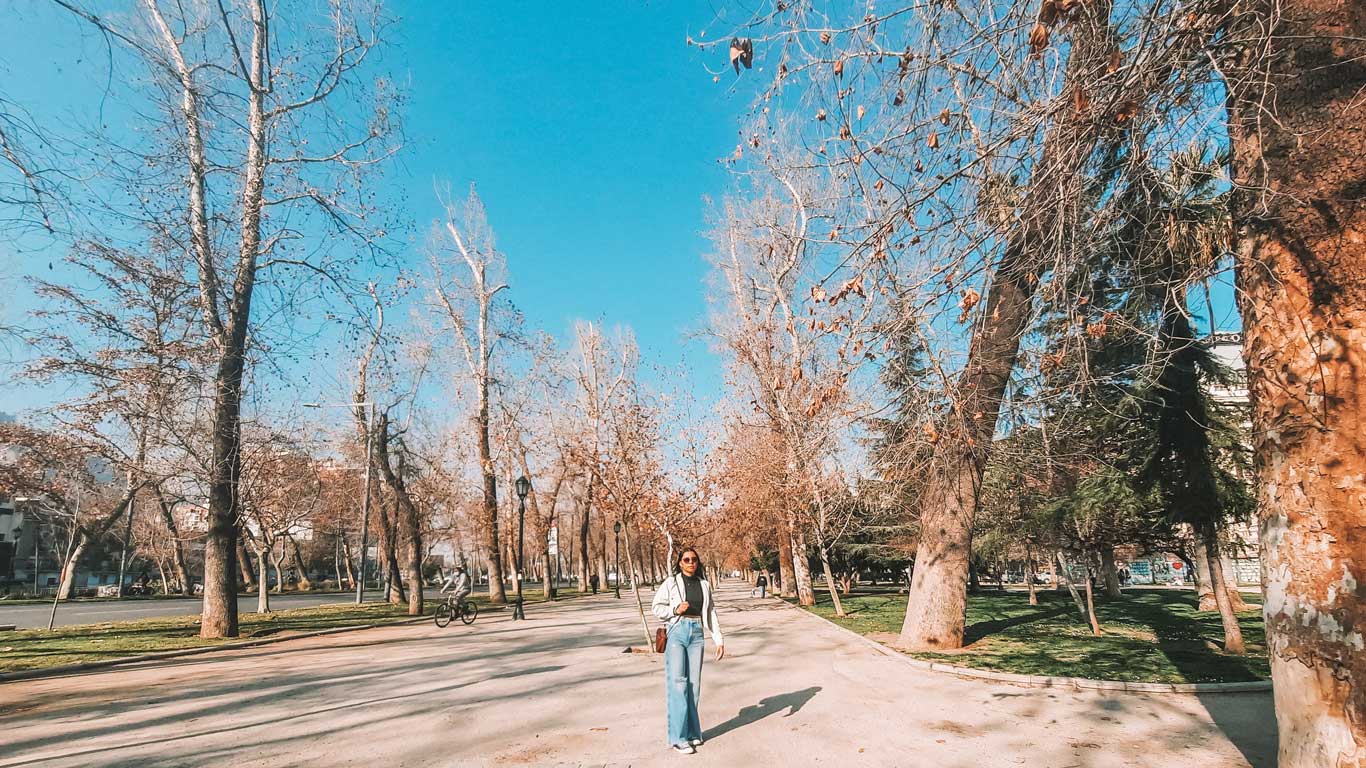 Una mujer caminando en un camino del Parque Forestal en Santiago de Chile, rodeada de árboles altos y delgados con hojas marrones de otoño. El cielo está despejado y azul, y al fondo se ven más personas disfrutando del parque.