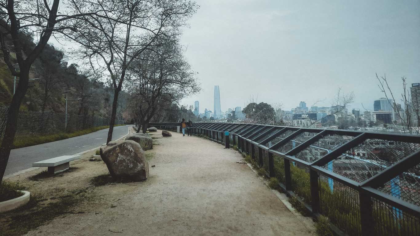 Camino en el Cerro San Cristóbal con vistas a la ciudad de Santiago al fondo. El cielo está nublado, y algunas personas caminan por la senda rodeada de árboles desnudos y grandes rocas. Al fondo se destaca el rascacielos Costanera Center.