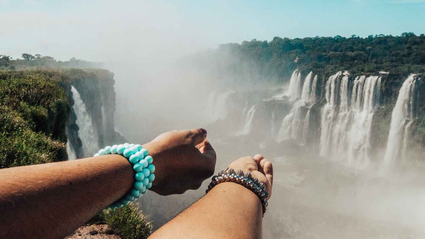 Primer plano de los brazos de una persona extendidos hacia las Cataratas del Iguazú en Argentina, con brazaletes coloridos en sus muñecas. Al fondo, se observan las imponentes cascadas rodeadas de vegetación y una ligera neblina creada por el agua en caída libre.