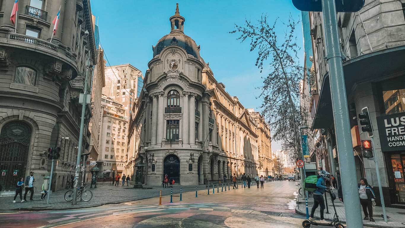 Una vista de una calle en el centro de Santiago de Chile, mostrando edificios históricos y personas caminando. La arquitectura clásica del edificio principal, con su cúpula y detalles ornamentales, resalta bajo un cielo azul. Ideal opción de dónde alojarse en Santiago de Chile, este barrio combina historia y modernidad.