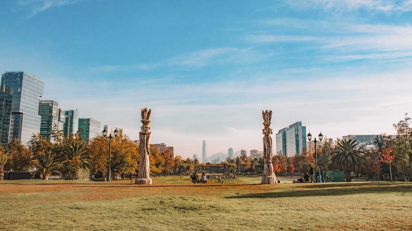 Vista de un parque en Las Condes, Santiago de Chile, con dos esculturas de madera en el centro. Al fondo, se pueden ver modernos edificios y el rascacielos Costanera Center bajo un cielo azul claro. La combinación de arte, naturaleza y arquitectura urbana destaca la diversidad del barrio.