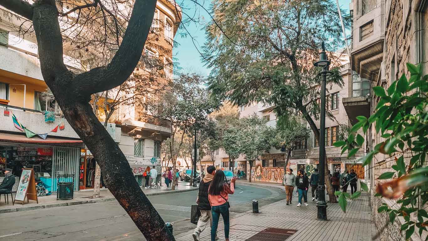 Imagen de una calle del Barrio Lastarria en Santiago de Chile, con personas caminando y árboles que bordean el camino. A la izquierda, hay una tienda con decoración colorida y un cartel en la acera. Los edificios a ambos lados de la calle tienen un diseño clásico y están parcialmente cubiertos por árboles.