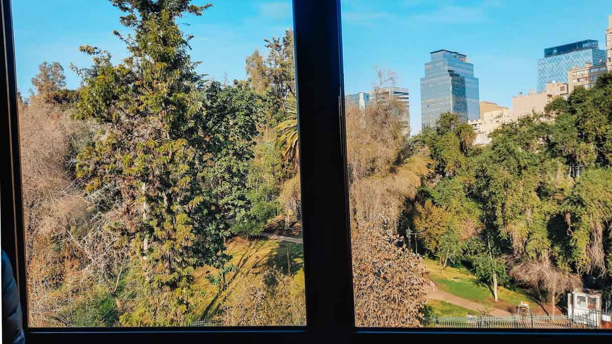 Vista desde una ventana del Foresta Hotel en Santiago de Chile, mostrando el Cerro Santa Lucía con diversos árboles y edificios modernos al fondo bajo un cielo azul claro. La mezcla de naturaleza y arquitectura urbana resalta en esta escena.