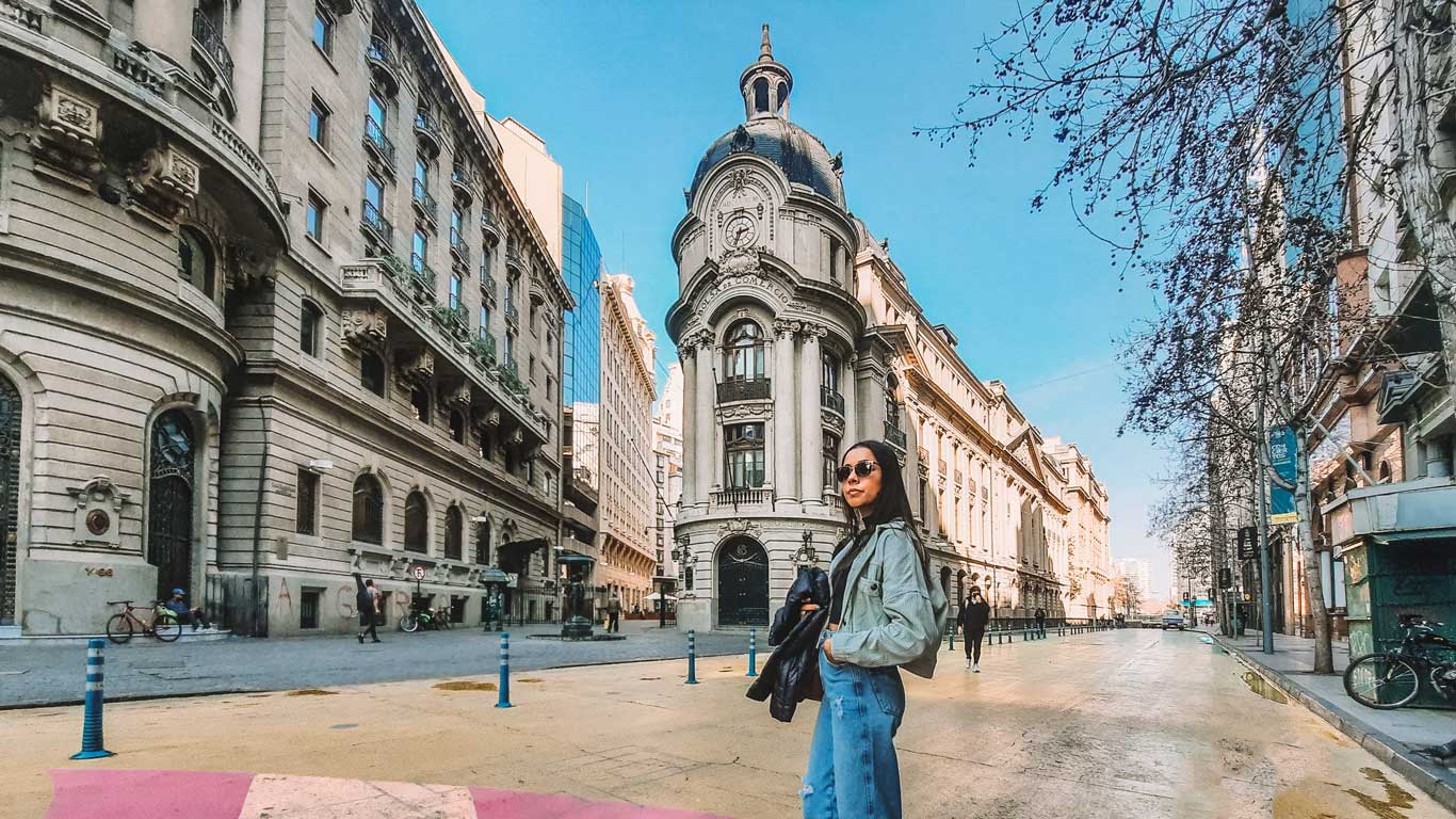 Una mujer con gafas de sol está posando en una calle del centro de Santiago de Chile, con edificios históricos y arquitectura neoclásica a su alrededor.