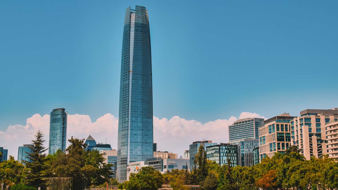 Vista de rascacielos modernos en Providencia, Santiago de Chile, destacando el icónico Costanera Center bajo un cielo azul despejado. La vegetación en primer plano contrasta con la arquitectura contemporánea de los edificios.