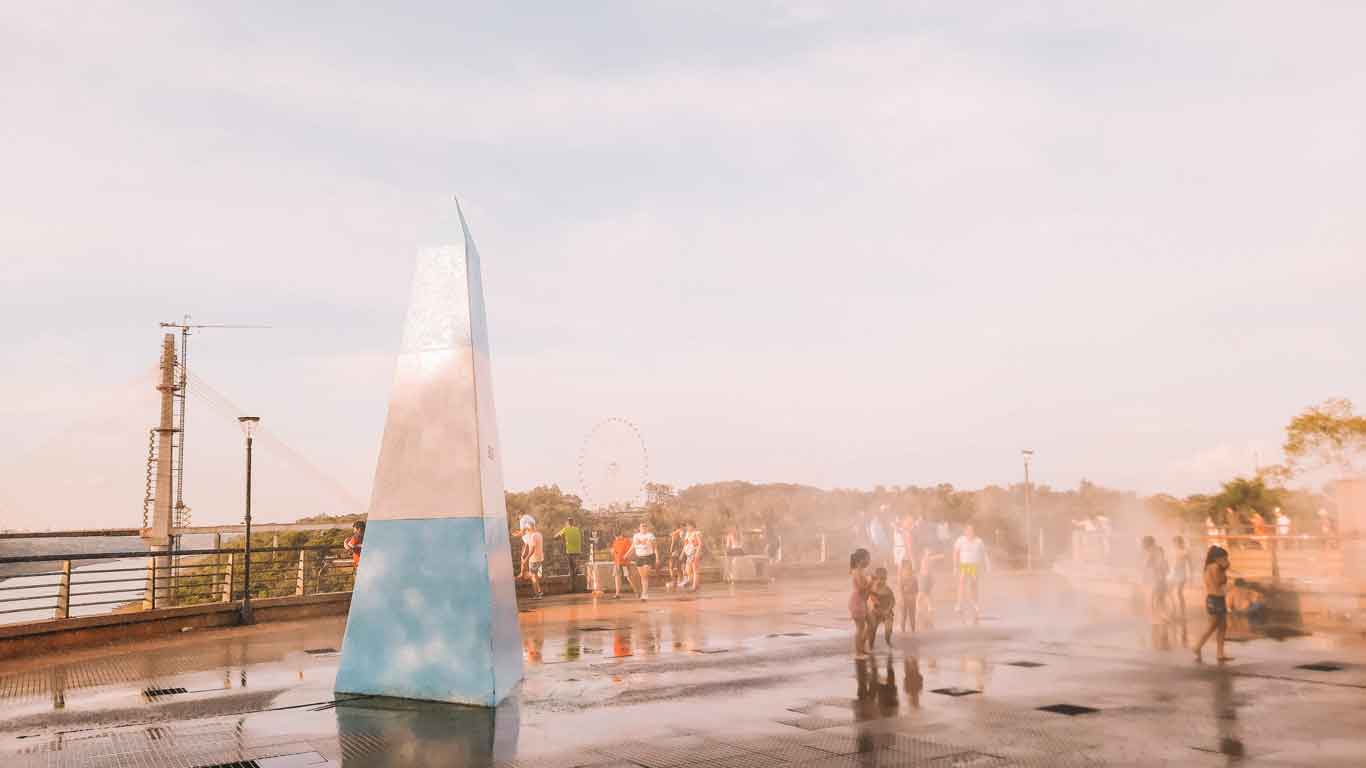 Hito de las Tres Fronteras en Puerto Iguazú durante el atardecer, con niños jugando en el agua que brota de la fuente.