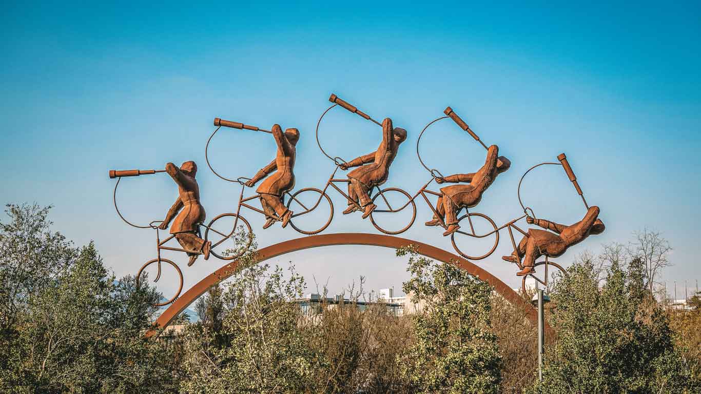 Escultura en Vitacura, Santiago, que muestra figuras montadas en bicicletas con trompetas, situada sobre un arco. La obra de arte se destaca contra un cielo azul claro, rodeada de árboles verdes, simbolizando el dinamismo y la creatividad del barrio.