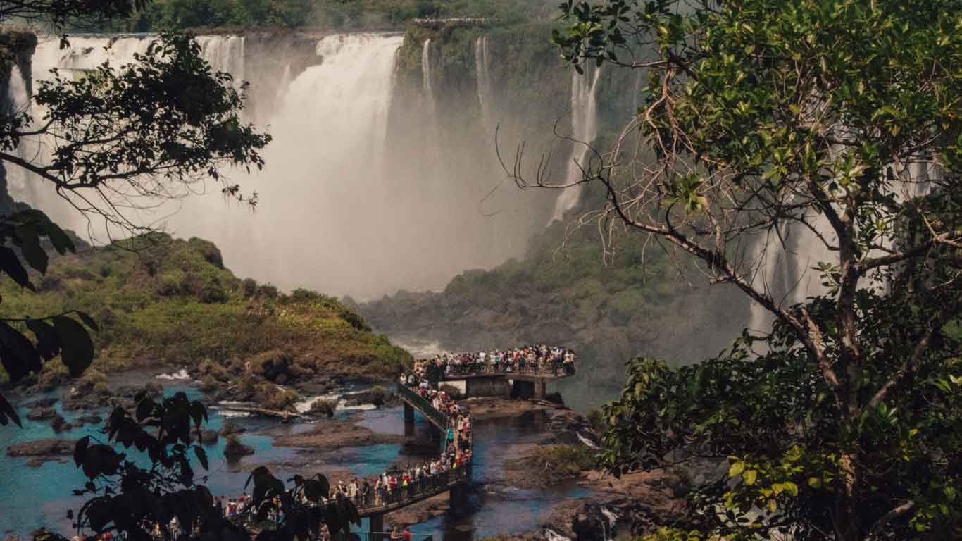 Una imagen de las cataratas del Iguazú, donde una multitud de visitantes se encuentra en una pasarela observando las majestuosas cascadas que se desploman entre la vegetación exuberante. La escena está enmarcada por árboles y ramas, mostrando la impresionante belleza natural de las cataratas del Iguazú.