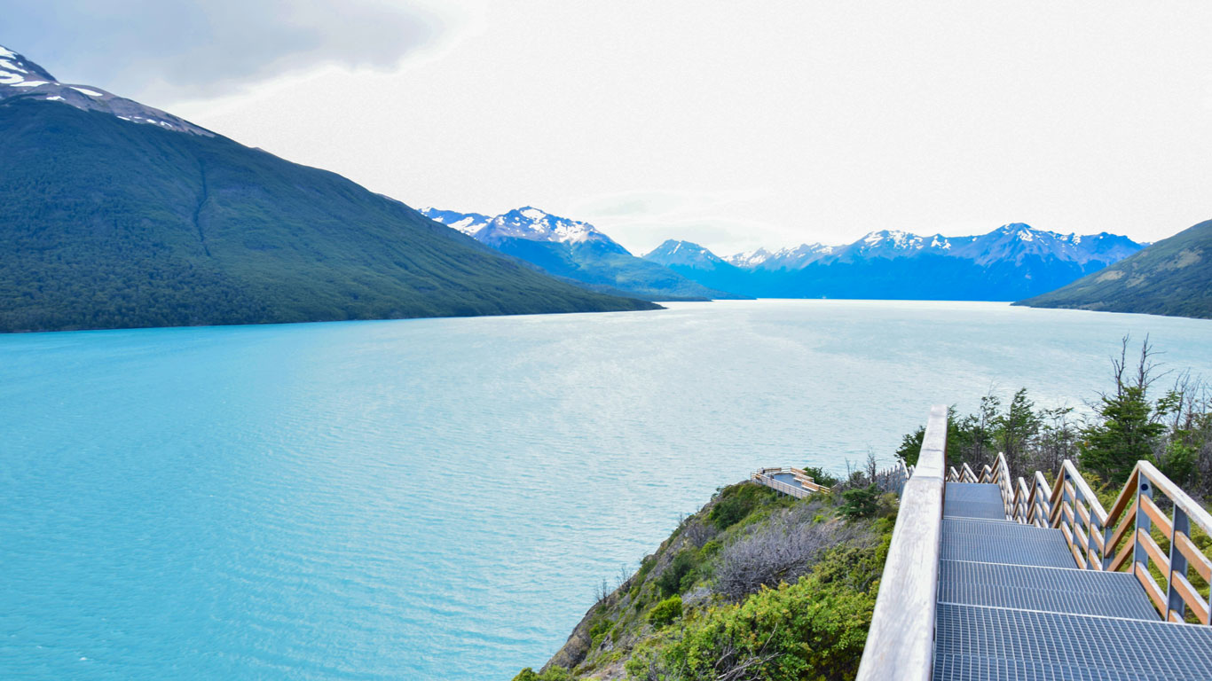 Una pasarela de madera, bordeando un lago con aguas azules y rodeado de montañas nevadas, en El Calafate, en Argentina.