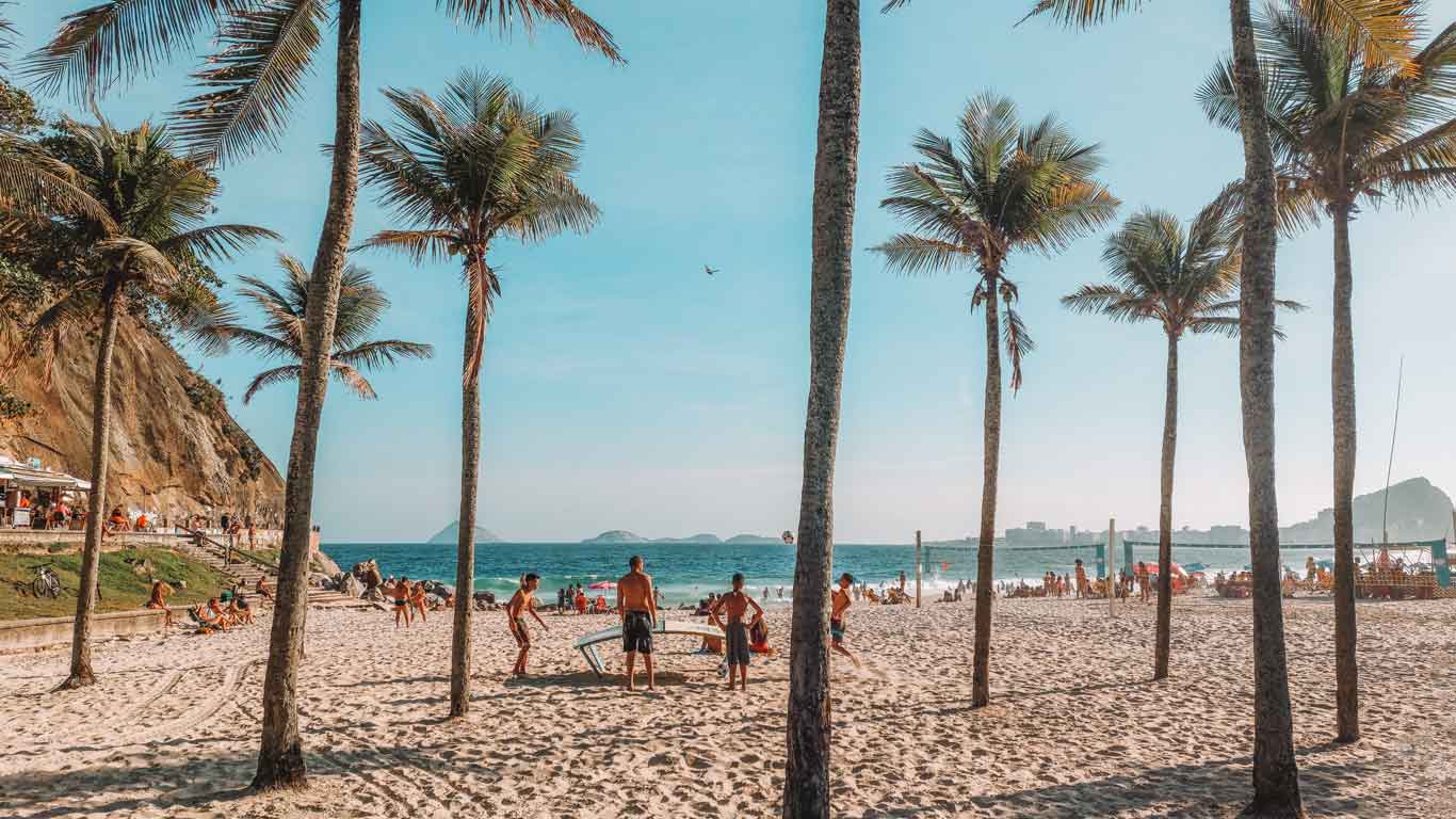 Playa de Leme en Río de Janeiro, con personas disfrutando del día soleado entre palmeras y arena clara. Grupos juegan al voleibol y frescobol, mientras otros se relajan a la sombra. El mar azul y el cielo despejado completan el escenario vibrante y animado.