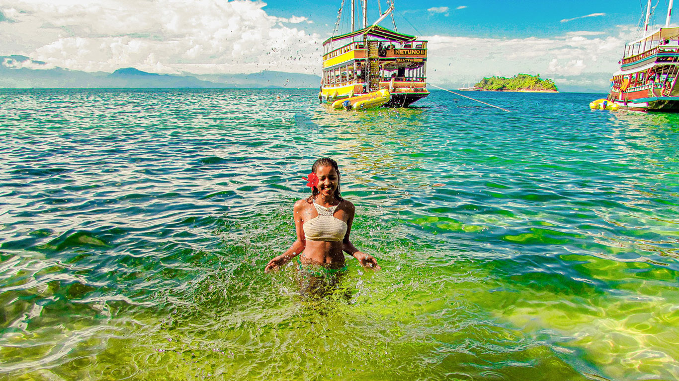 Mujer con una flor roja en el cabello lanzando agua hacia arriba con las manos en una playa de Paraty, de aguas verdes. Al fondo, una goleta se mezcla en el horizonte con el cielo azul.