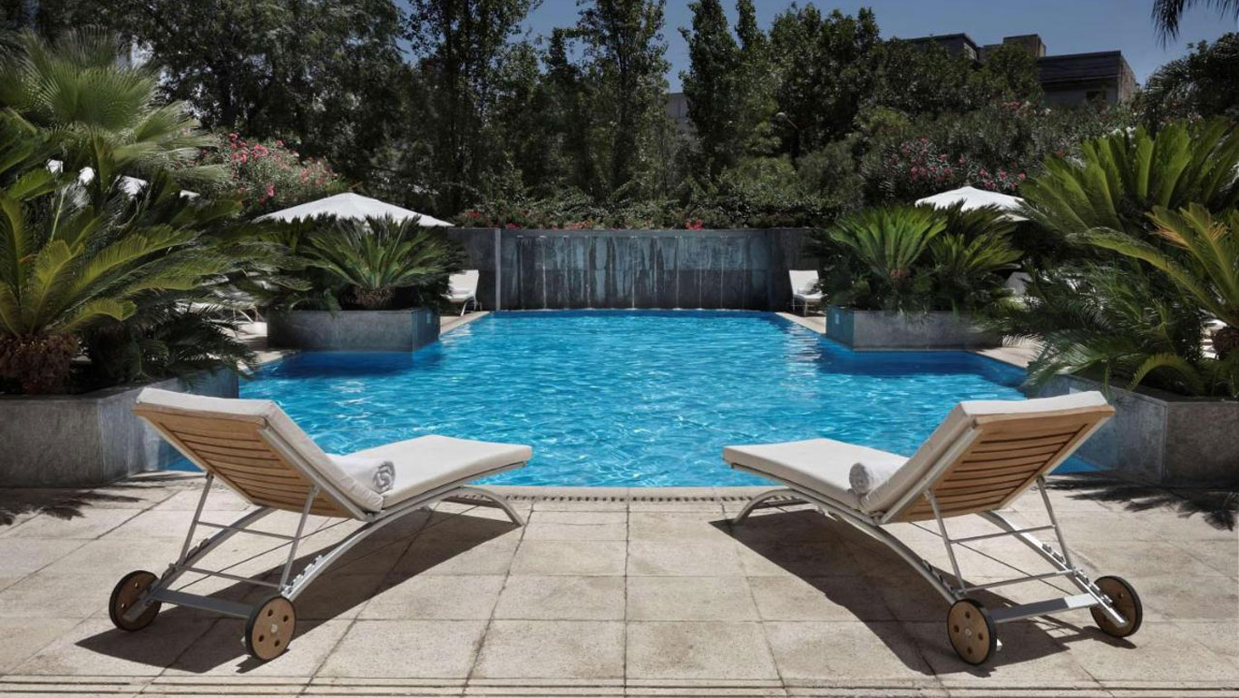 La imagen muestra una elegante piscina al aire libre rodeada de vegetación exuberante en el Park Hyatt Mendoza. Dos tumbonas con toallas enrolladas están dispuestas al borde de la piscina, invitando al relax bajo el sol. Las plantas tropicales y el entorno natural crean un ambiente tranquilo y lujoso.