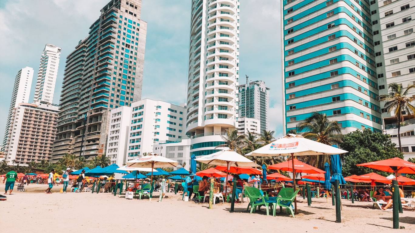 Vista de la playa en Bocagrande, Cartagena de Indias, con altos edificios modernos al fondo y una colorida escena de sombrillas rojas y azules en la arena. Los turistas disfrutan del día soleado en la playa, rodeados de palmeras y el vibrante ambiente costero característico de esta popular zona turística.