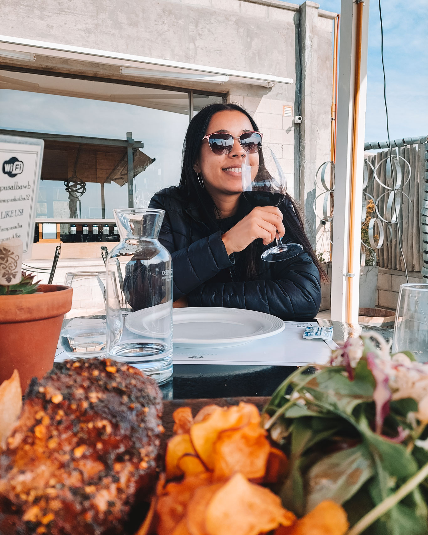 La imagen muestra a una mujer disfrutando de una copa de vino tinto en una mesa al aire libre, en lo que parece ser un restaurante de una bodega. Lleva gafas de sol y una chaqueta, mientras sonríe y se relaja. En primer plano, se observa un plato de comida con papas fritas y ensalada, complementando la experiencia gastronómica. El entorno sugiere un ambiente acogedor y tranquilo.