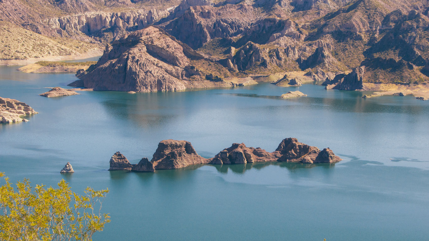 La imagen muestra el Cañón del Atuel en Mendoza, Argentina, con formaciones rocosas sobresaliendo de las aguas cristalinas de un embalse. Las montañas áridas y rocosas rodean el cuerpo de agua, creando un paisaje imponente y sereno. La vegetación escasa en primer plano resalta la belleza natural del lugar.