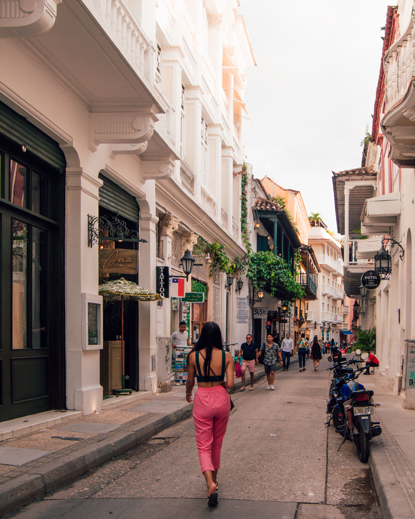 Una mujer camina por una pintoresca calle de la Ciudad Vieja en Cartagena de Indias, rodeada de edificios coloniales de fachadas blancas con balcones adornados con vegetación. A los lados de la calle, tiendas locales y restaurantes crean un ambiente vibrante mientras peatones y motos llenan el espacio. La escena captura la vida cotidiana y el encanto histórico de este barrio emblemático.