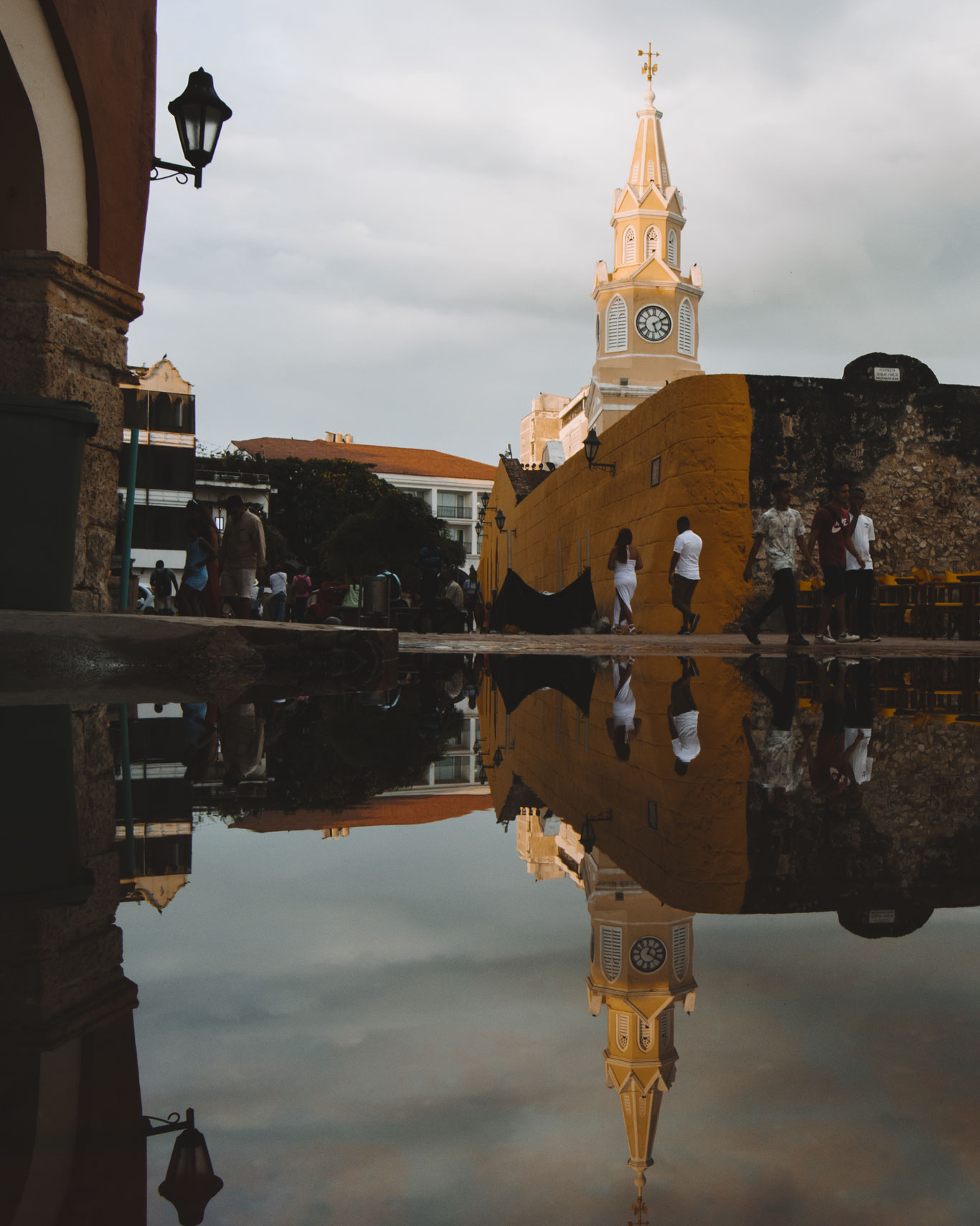 Una vista icónica de la Torre del Reloj en la Ciudad Vieja de Cartagena de Indias, reflejada en un charco tras una lluvia. La estructura amarilla se alza sobre la muralla histórica, mientras varios peatones caminan por la plaza. La escena captura la esencia colonial y el encanto atemporal de este sitio emblemático de Cartagena de Indias.