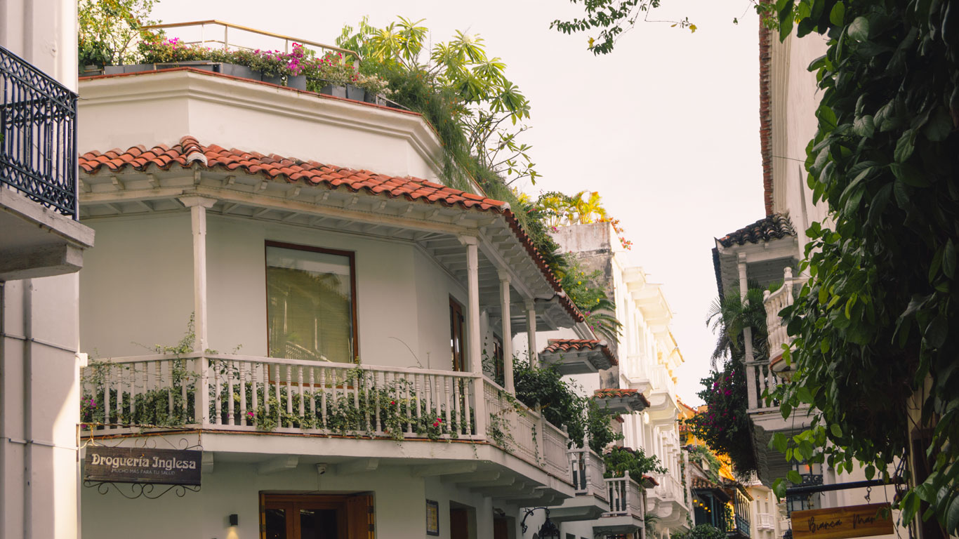 Una vista de una calle colonial en Cartagena de Indias, con casas de dos pisos que presentan balcones adornados con plantas y flores. Los tejados de tejas rojas y las paredes blancas son característicos de la arquitectura tradicional de la ciudad. En la esquina, se puede ver un letrero que dice "Droguería Inglesa", añadiendo un toque local a la escena.