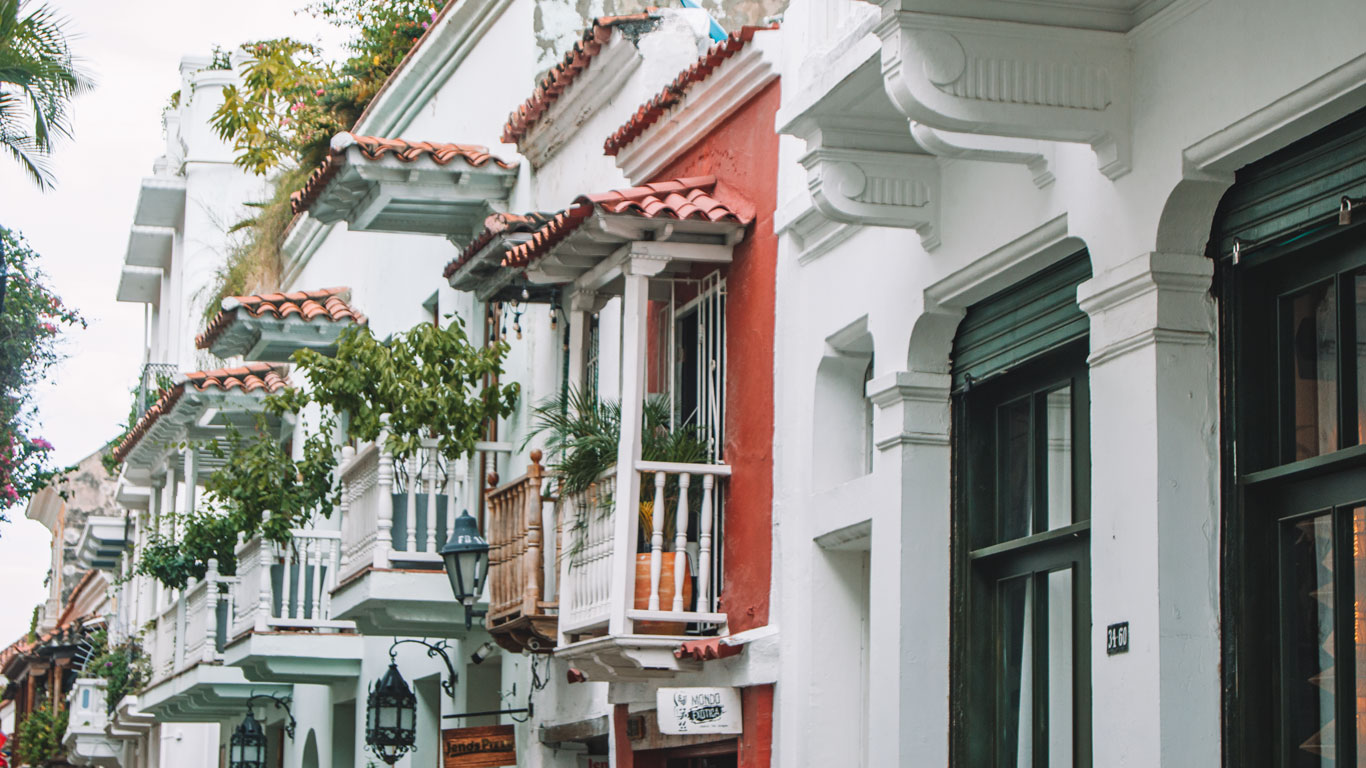 Una colorida fila de casas coloniales en Cartagena de Indias, con balcones de madera y plantas colgantes que decoran las fachadas. Las paredes blancas y los tejados de tejas rojas reflejan el encanto arquitectónico de la ciudad. Un lugar emblemático de la zona histórica de Cartagena de Indias.