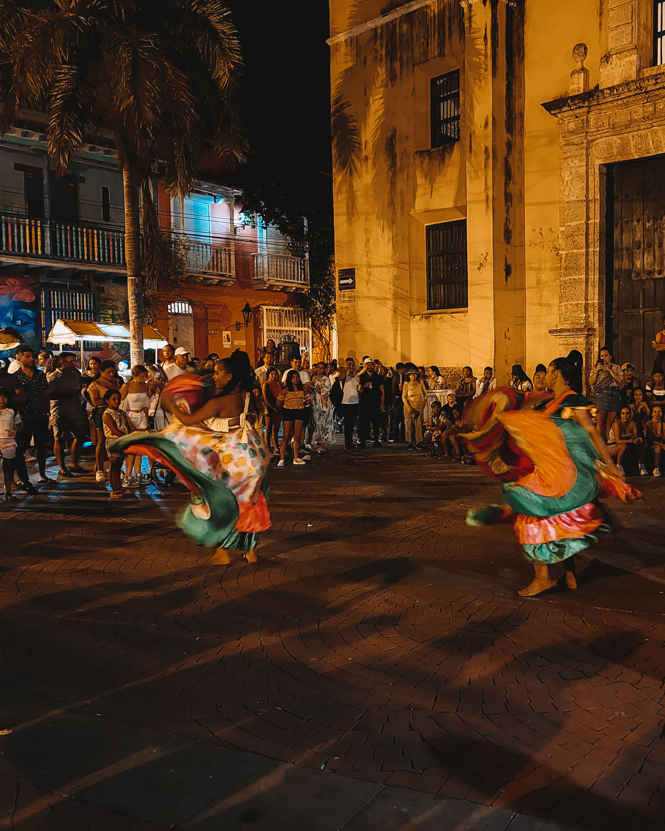 Un vibrante espectáculo nocturno en las calles de Cartagena de Indias, donde dos mujeres bailan con coloridos trajes tradicionales frente a un público entusiasta. El ambiente festivo está enmarcado por la arquitectura colonial y las palmeras típicas de la ciudad, capturando la energía cultural de la noche en el barrio de Getsemaní.