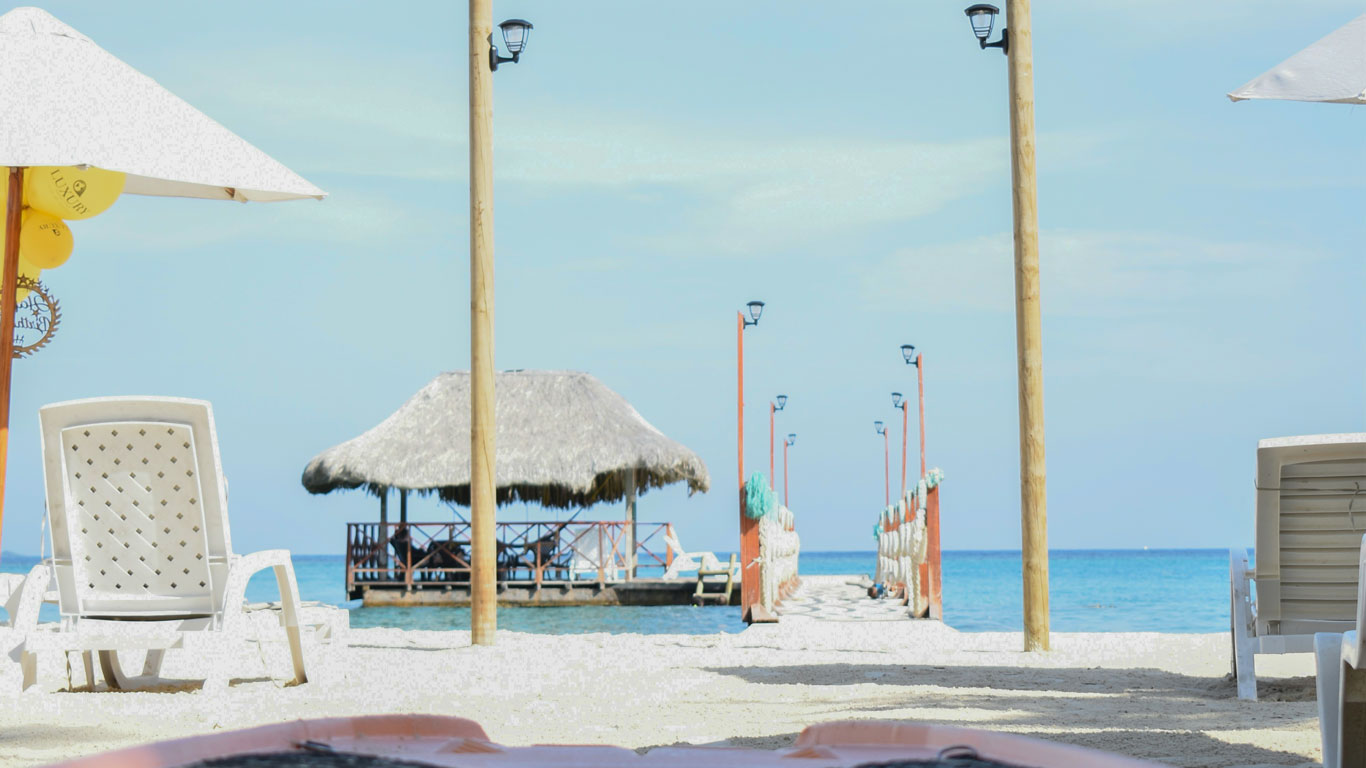 Una playa tranquila en las Islas del Rosario, cerca de Cartagena de Indias, con sillas y sombrillas blancas frente a un muelle de madera que lleva a un quiosco cubierto de paja sobre el agua cristalina. El cielo despejado y el mar azul crean un ambiente sereno y relajante, ideal para disfrutar de la belleza natural de este paraíso tropical.