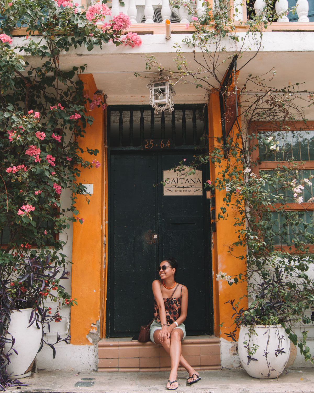 Una mujer sentada en la entrada de una casa colonial en Cartagena de Indias, rodeada de coloridas flores y vegetación trepadora. La fachada de la casa presenta paredes de color naranja y detalles florales, con un letrero que dice “Casa Gaitana”. Esta escena refleja el encanto relajado y vibrante de la ciudad.