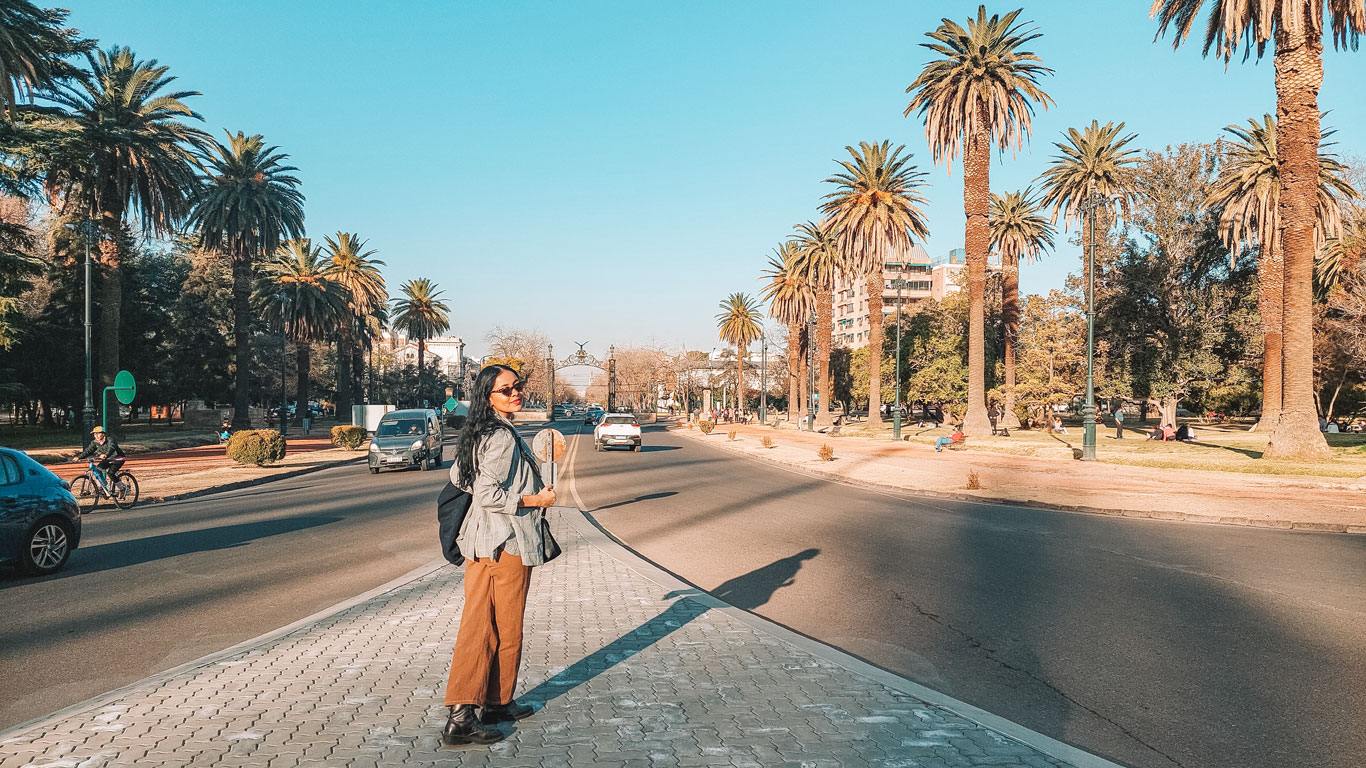 Mujer caminando frente a la entrada del Parque General San Martín en Mendoza. La mujer, de cabello largo y oscuro, lleva pantalones vaqueros marrones, un blazer gris y gafas de sol oscuras.