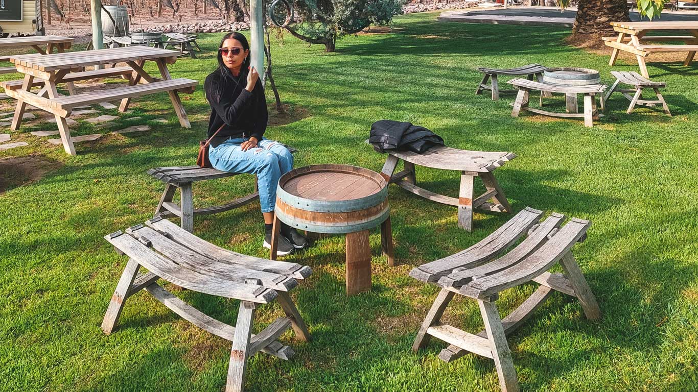 Mujer sentada en una zona verde de la Bodega Tempus Alba en Mendoza, rodeada de bancos de madera rústicos y césped verde, en un ambiente relajante de viñedo.