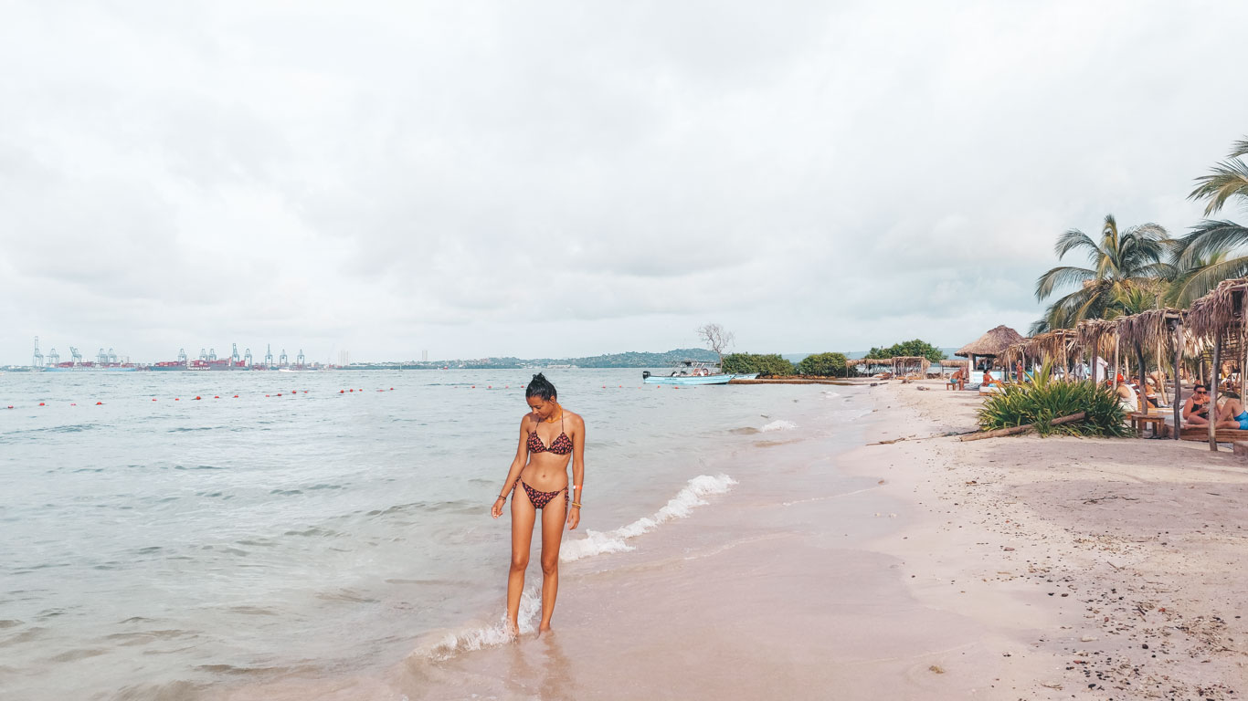 Una mujer camina por la orilla de la playa en Tierra Bomba, cerca de Cartagena de Indias, disfrutando de un día tranquilo junto al mar. A lo lejos, se puede ver el puerto industrial en contraste con las palmeras y cabañas rústicas que bordean la playa. El ambiente relajado invita a desconectarse en esta isla cercana a la ciudad.