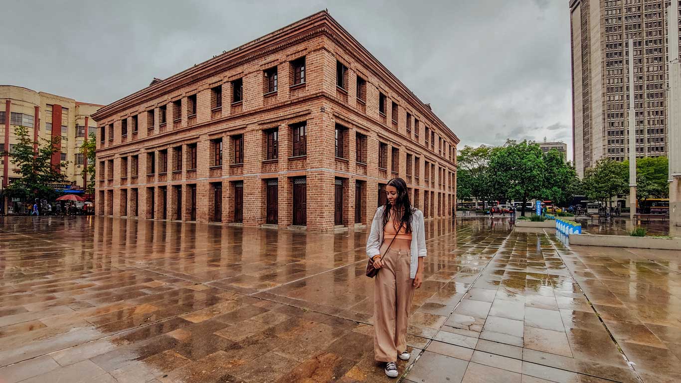 Una escena en el barrio La Candelaria, Medellín, donde se destaca un edificio de ladrillo de estilo clásico bajo un cielo nublado. El suelo de la plaza está mojado, reflejando la reciente lluvia, y una persona camina en primer plano vestida con ropa casual. Al fondo, se observan árboles, edificios modernos y una zona de actividad urbana con algunas personas y estructuras comerciales.