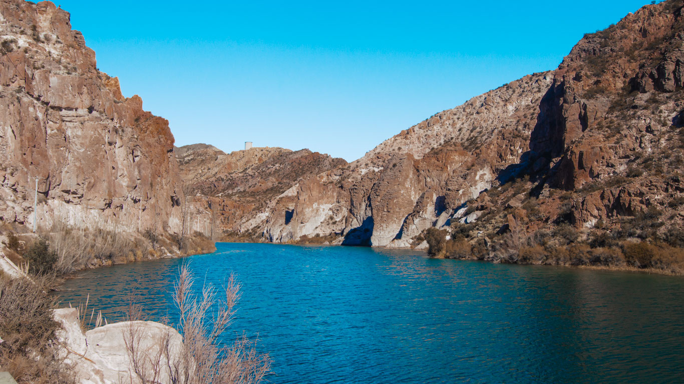 Esta imagen muestra el Cañón del Atuel en San Rafael, Mendoza. Se pueden ver imponentes paredes rocosas que flanquean un río de aguas azules, en un paisaje árido y montañoso bajo un cielo despejado.