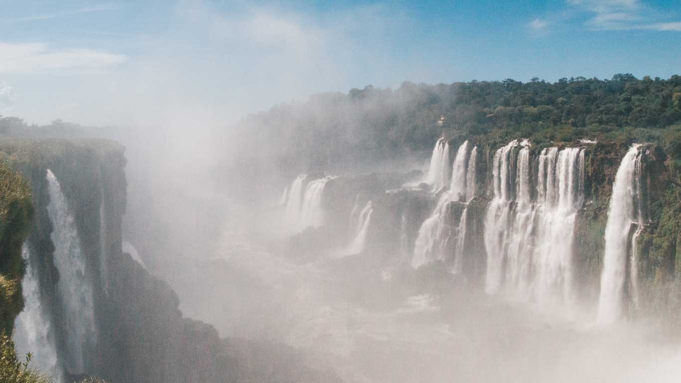 La imagen muestra una vista panorámica de las impresionantes Cataratas del Iguazú, con múltiples cascadas cayendo desde altas formaciones rocosas. El agua genera una niebla espesa que cubre parcialmente la escena, mientras la vegetación densa rodea las caídas. El cielo despejado resalta la majestuosidad del paisaje, que es uno de los atractivos naturales más impactantes del mundo.