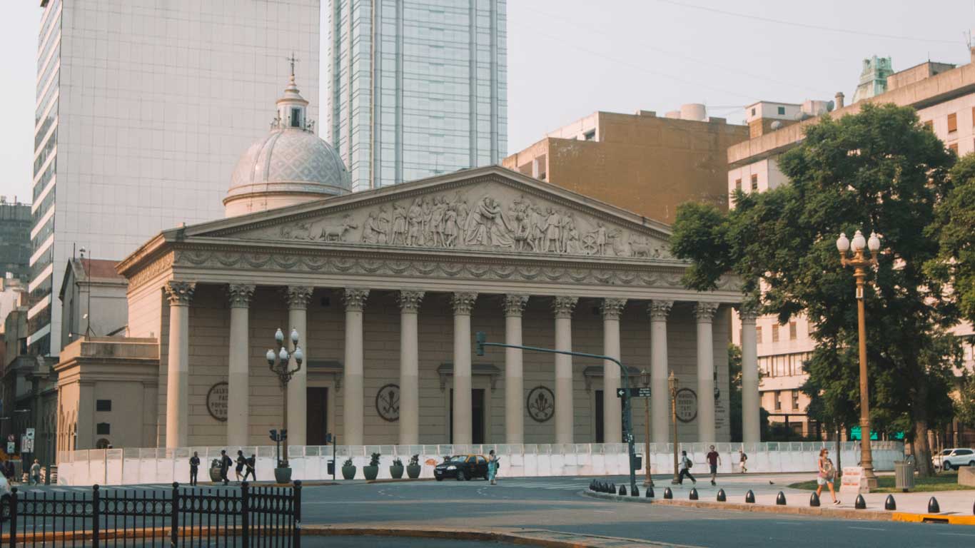 Imagen de la Catedral Metropolitana de Buenos Aires, Argentina, con su fachada neoclásica adornada por columnas y un frontón con relieves escultóricos. Se observa la cúpula detrás de la estructura principal, rodeada por edificios modernos. El entorno muestra una calle tranquila con algunas personas caminando y árboles en el lado derecho de la imagen.