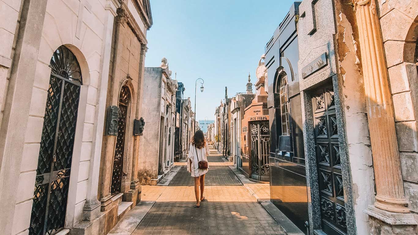 Una persona caminando por un estrecho pasillo del Cementerio de la Recoleta en Buenos Aires, Argentina. A ambos lados se observan mausoleos ornamentados y tumbas de diversos estilos arquitectónicos, bajo un cielo despejado. El ambiente es tranquilo y solemne, realzando la atmósfera histórica del lugar.