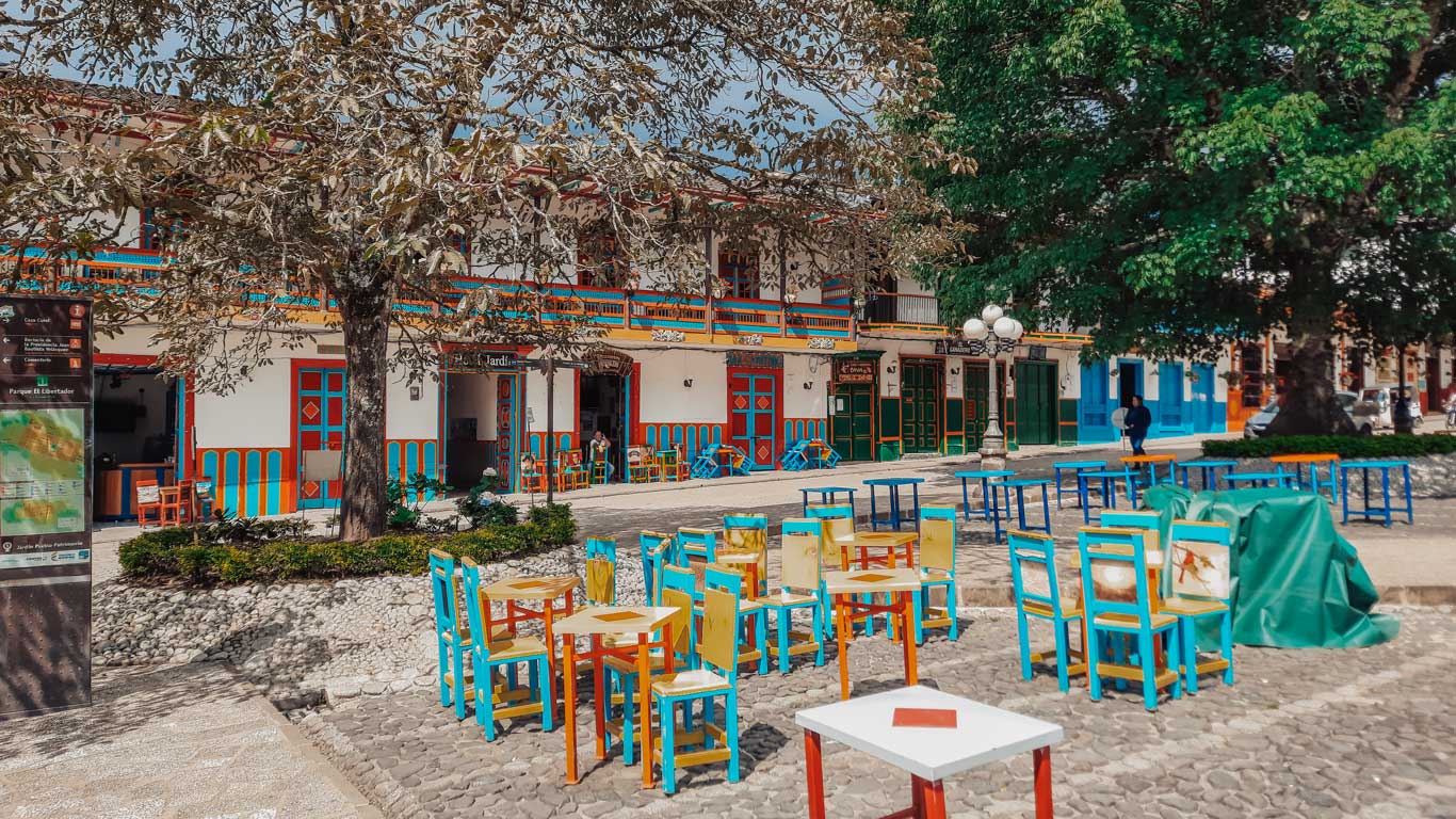 Un colorido café al aire libre en la plaza principal de Jardín, Antioquia, con sillas y mesas pintadas en tonos brillantes de azul, amarillo y rojo. Al fondo, se observa una fachada tradicional con balcones decorados y el "Hotel Jardín". Grandes árboles proporcionan sombra, creando un ambiente acogedor y relajado en el centro del pueblo, mientras algunas personas disfrutan del entorno pintoresco.