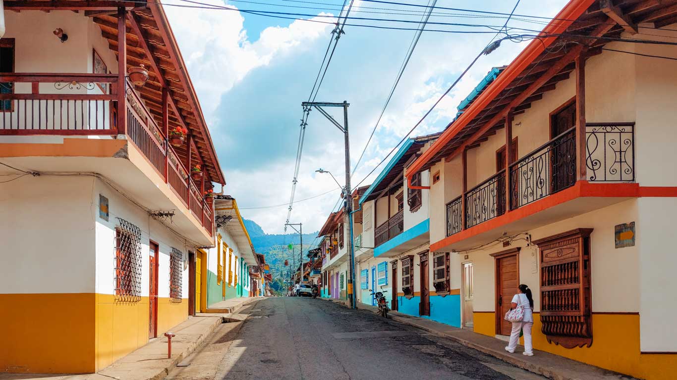 Una calle tranquila en Jardín, Colombia, con casas de estilo tradicional pintadas en colores vibrantes, como blanco, amarillo y azul, destacando los balcones de madera con detalles ornamentales. Una persona vestida de blanco camina por la acera, mientras el cielo despejado y las montañas al fondo completan la escena. Esta imagen refleja el encanto del pueblo, perfecto para quienes buscan donde alojarse en Jardín Colombia, rodeados de un ambiente pintoresco y acogedor.